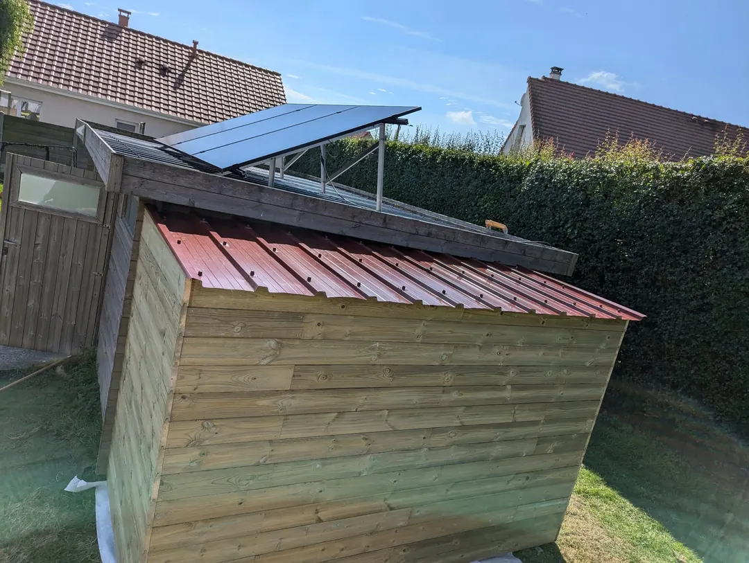 Abri de jardin en bardage bois horizontal avec toit en tôle nervurée rouge, adossé à un abri existant.