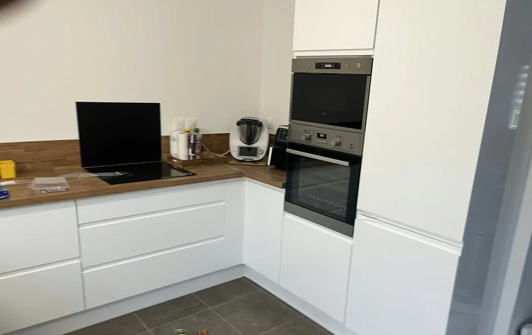 Cuisine blanche laquée sans poignées, plan de travail en bois, colonne four et micro-ondes, carrelage gris.