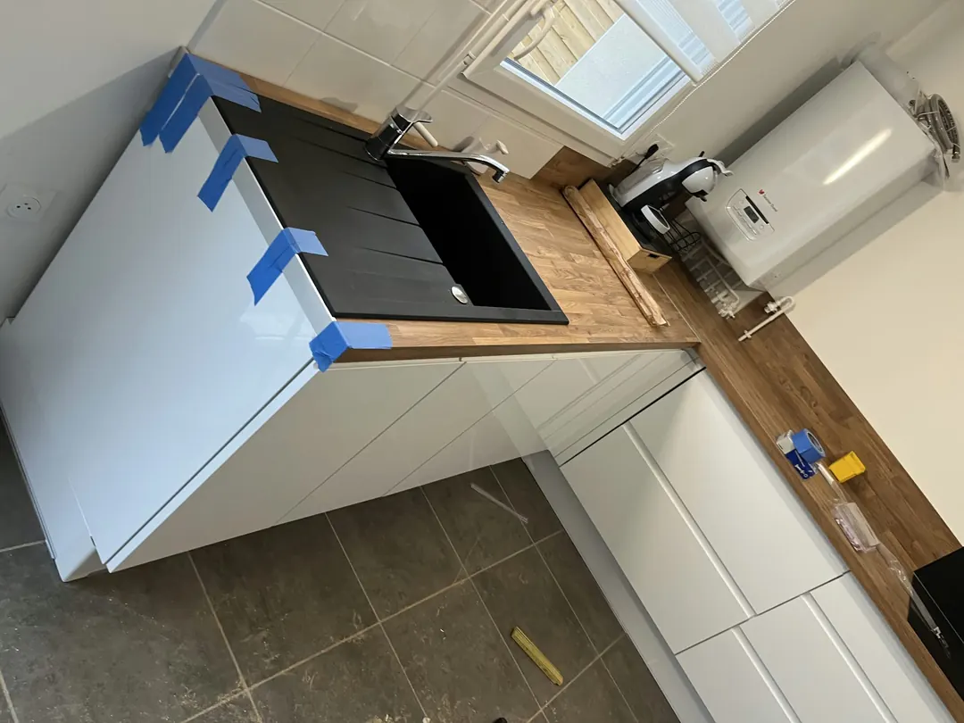 Meubles de cuisine blancs, plan de travail en bois avec évier noir encastré, adhésif bleu de maintien.