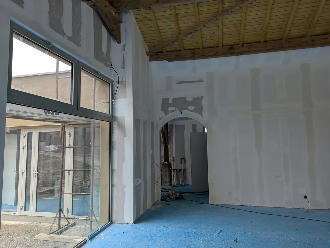 Grande pièce en travaux : baie vitrée haute, murs en plaques de plâtre avec bandes, passage en arche, charpente en bois au plafond.