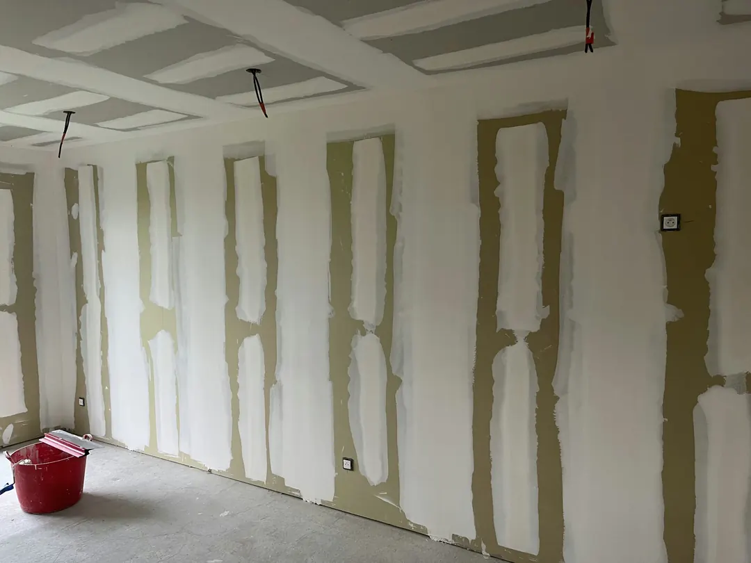 Murs en plaques de plâtre avec les joints enduits en blanc, plafond aux joints enduits, seau rouge au sol.