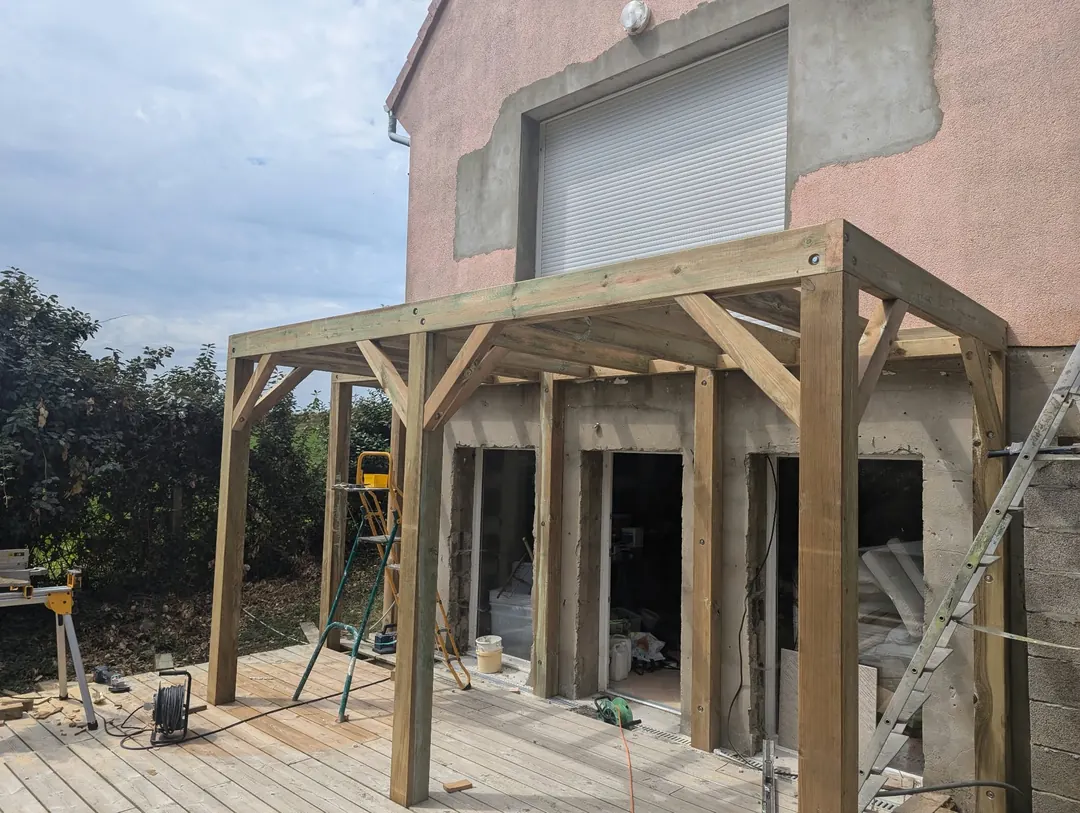 Structure de pergola en bois (poteaux, jambes de force, poutres) montée sur une terrasse devant une maison.