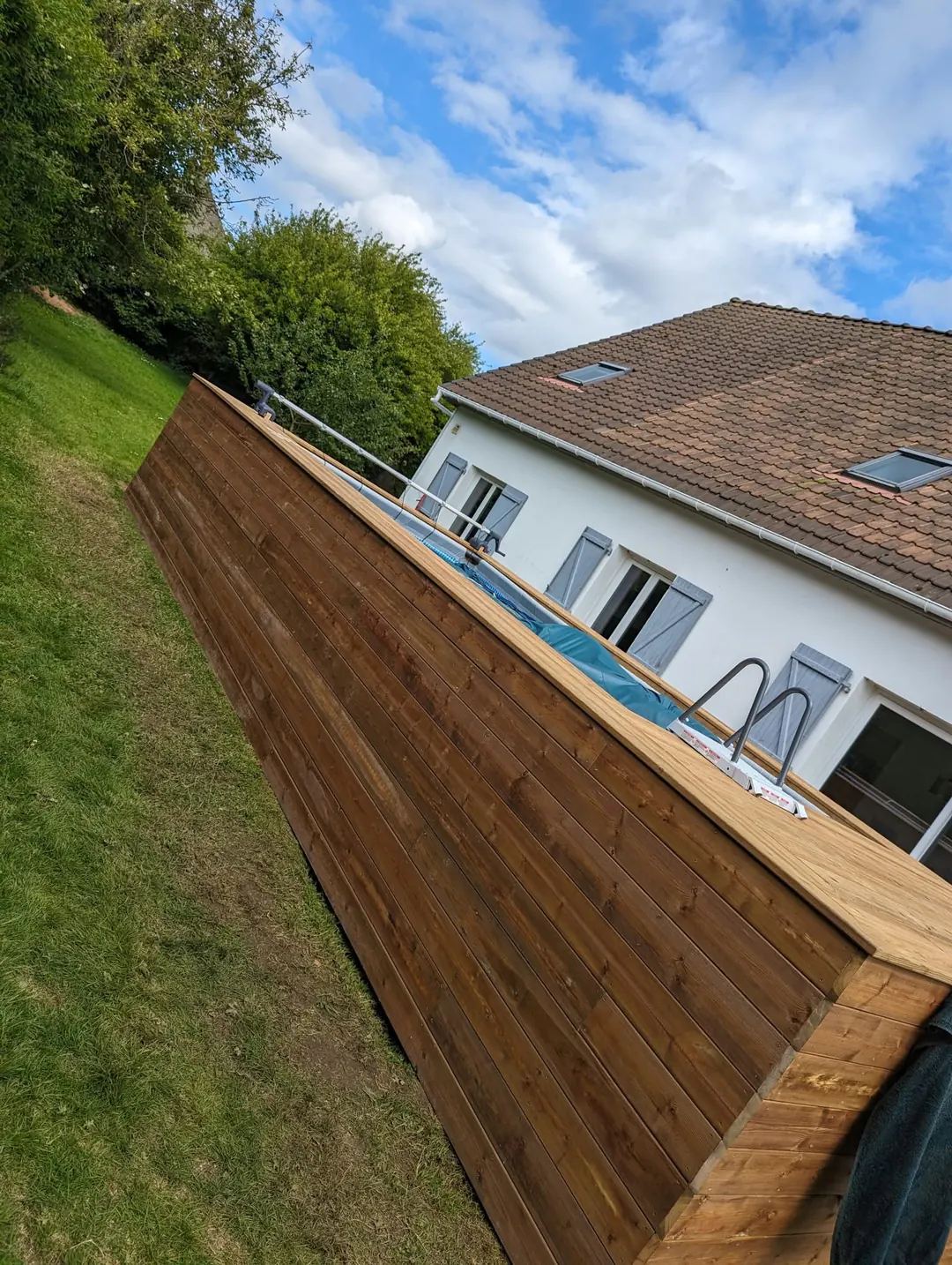 Long côté d'une piscine hors-sol habillée de lames de bois, maison blanche au fond.