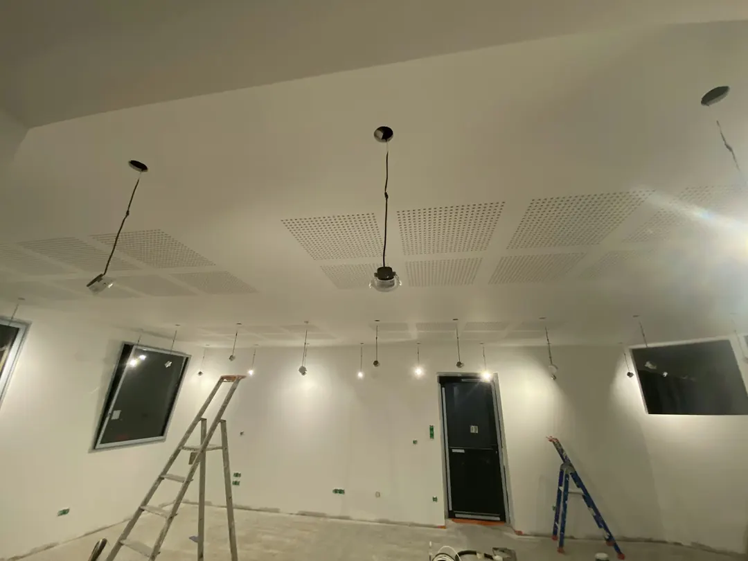 Pièce blanche avec un plafond en plaques perforées, spots encastrés allumés, escabeaux au sol.