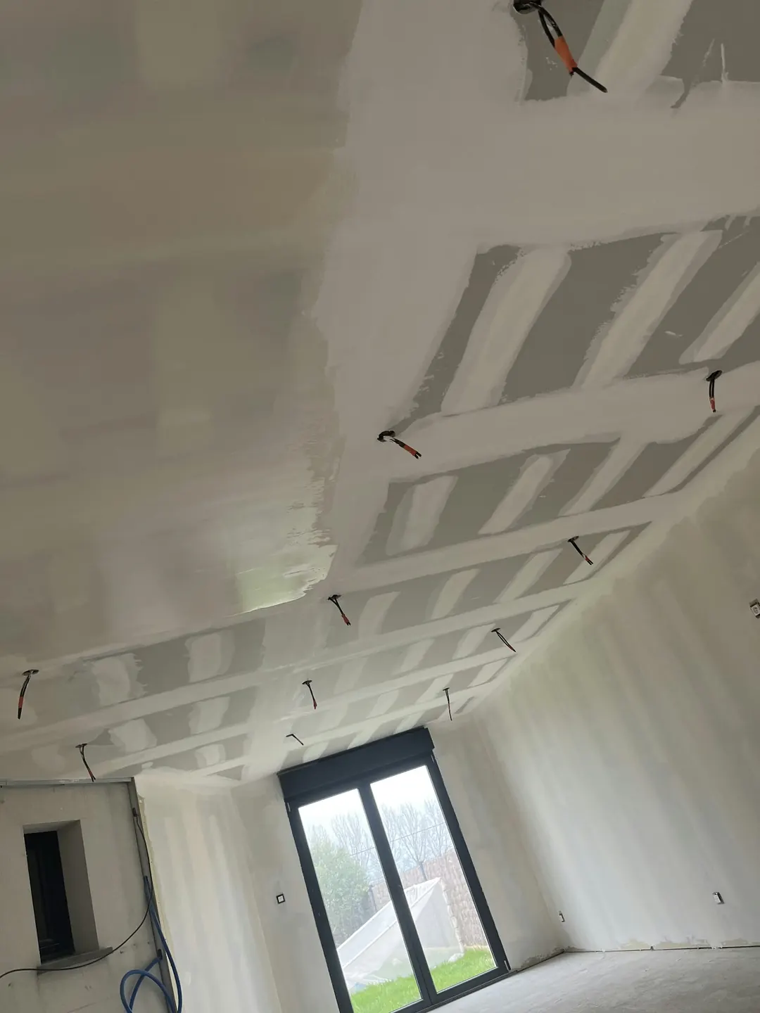 Plafond en plaques de plâtre aux joints enduits, fils électriques en attente, porte-fenêtre au fond.