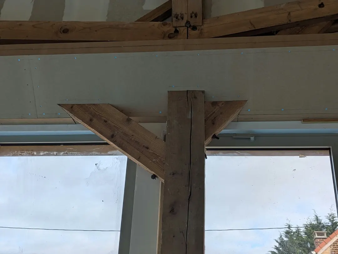 Poteau en bois ancien et ses deux jambes de force devant un mur en plaques de plâtre et une grande fenêtre.