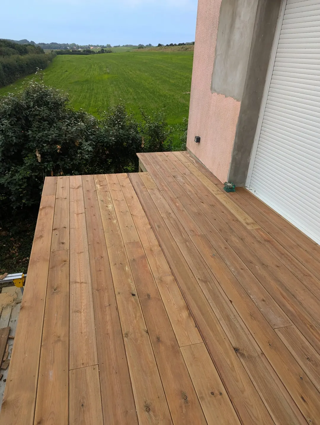 Terrasse neuve en lames de pin le long d'une maison, vue sur une prairie et des haies.