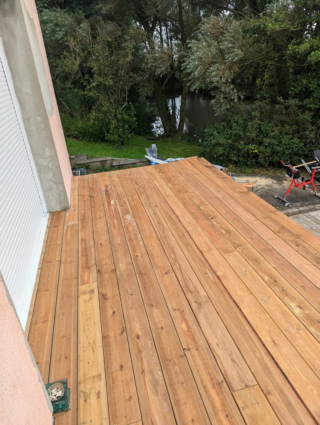 Lames de terrasse en pin fraîchement posées le long d'une façade, jardin arboré au fond.