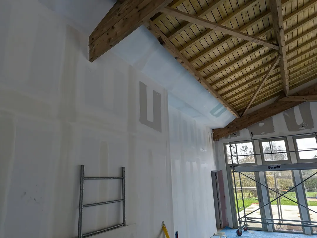 Mur haut en plaques de plâtre aux joints enduits, sous une charpente en bois apparente et un lambris.
