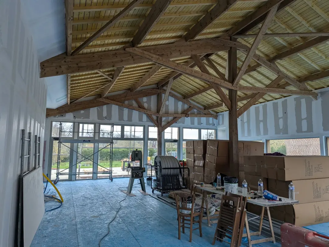 Très grande pièce sous des fermes de charpente en bois apparentes, murs en plaques de plâtre, cartons et outils au sol.
